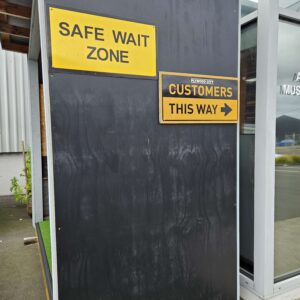 Black painted hoarding ply outside with a yellow sign reading “SAFE WAIT ZONE” and another yellow sign reading “CUSTOMERS THIS WAY.”